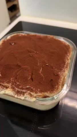 A close-up, eye-level shot shows a rectangular glass dish filled with a layered dessert, likely tiramisu, dusted generously with cocoa powder. The dessert occupies the majority of the frame, set against a dark, reflective surface that appears to be a modern stovetop with subtle circular markings. The dessert itself is creamy and white, with visible layers of ladyfingers or cake beneath the cocoa topping. The texture of the cocoa powder is uneven, with some areas more densely dusted than others, creating a rustic appearance. The glass dish has slightly rounded corners and a thin rim. In the background, out of focus, a blurred white surface and a hint of a shelf or cabinet suggest an indoor setting, possibly a kitchen. The lighting is soft and appears to be coming from above and to the side, illuminating the dessert and highlighting the sheen on the stovetop. There is a small, dark, irregular shape on the stovetop in the foreground, perhaps a spill or a reflection. The overall impression is one of a freshly prepared dessert, ready to be served. Visible text on the stovetop reads "INDUCTION". The scene is static, with no discernible activity other than the presentation of the dessert. The time of day and weather are not apparent. The location context is Riihimäki, Finland, which is not explicitly indicated in the visual but provided as context.
