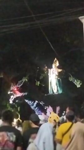 A nighttime outdoor event is underway, with a large crowd of people gathered. In the background, a tall, illuminated figure stands above the crowd, holding a staff. In the foreground, a dragon-like effigy is being paraded through the crowd, emitting smoke and sparks. The effigy is dark with red accents on its head and appears to be made of fabric and possibly other materials. The crowd is diverse, with individuals of various ages and genders, many wearing casual clothing. The environment is dimly lit, with the primary illumination coming from the figures and potentially some ambient lighting from the surrounding area. Overhead, power lines are visible, indicating an urban or semi-urban setting. The atmosphere appears festive and energetic.