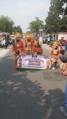 The video shows a parade of children in traditional Indonesian attire marching down a street. The children are wearing brightly colored outfits with gold headpieces and elaborate jewelry. They are holding a banner that reads "PAWAI BUDAYA NUSANTARA ADAT PALEMBANG TK AL HIDAYAH MANDESAN 01". The parade appears to be taking place on a sunny day, with trees and buildings lining the street. There are spectators on the side of the road, as well as other vehicles and motorbikes. The children are walking in a line, and some of them are smiling and waving. The overall atmosphere seems festive and celebratory.