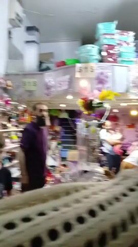 Four people are in a shop looking at items on a shelf. Three of the people are women wearing hijabs, and one is a young boy. The shelves are stocked with various household items including containers, kitchenware, and decorative objects. The women and the boy are examining items on the shelf, possibly considering a purchase. The shop appears to be brightly lit.
