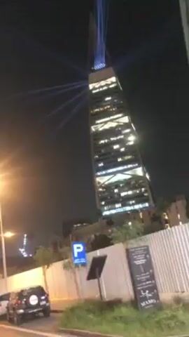 The video, shot at night, focuses on a tall building illuminated with blue laser beams emanating from its upper portion. The building appears to be a modern skyscraper with many lit windows. A street with a parked car and signage is visible in the foreground, though the camera's movement obscures it at times. The camera angle shifts slightly, but the building remains the primary subject throughout the video.