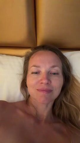 A woman lies in a bed with a light-colored pillow and a tan upholstered headboard behind her. The camera is positioned close to her face and the shot is taken from a low angle. She is looking at the camera with her eyes partially closed and a slight smile. Her hair is blonde and falls around her shoulders. The lighting in the room is warm and creates a soft glow. The video then pans upwards and moves quickly, becoming blurry.