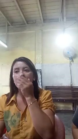 A woman in a yellow shirt with a floral pattern is sitting indoors. She is touching her face and hair. In the background, there are wooden benches and a fan. The lighting suggests it is daytime or artificial lighting is on.