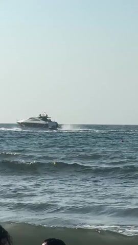 A yacht cruises across a choppy blue ocean under a clear sky. The yacht is white with dark accents and is leaving a white wake behind it. In the foreground, the tops of people's heads are visible, suggesting they are on a beach. The waves are rolling towards the shore.