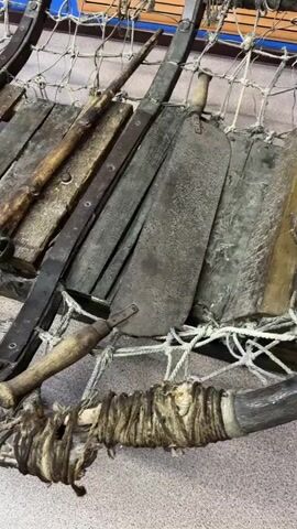 The video shows a close-up view of an old, rustic sled. The sled is constructed primarily of wood and metal, with a net-like material forming the bed. Several items are placed on the sled, including what appear to be two firearms, one longer and thinner than the other, and what might be a knife or machete with a wooden handle. The wood is weathered and appears aged, with visible grain and some damage. Metal straps and fasteners are used to hold the wooden slats together and attach them to the sled's frame. The sled is resting on a carpeted floor, and the background suggests an indoor setting, possibly a museum or historical display, with blue and wooden paneling visible. The lighting is somewhat dim, creating shadows that accentuate the textures and age of the objects.