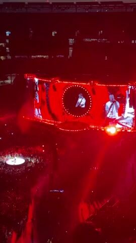 An aerial shot shows a large, crowded stadium during a concert. The stage is illuminated with bright red lights, and a massive screen displays images of a musician performing. The crowd, visible in the lower part of the frame, is also bathed in red light, with many people holding up their phones. The overall atmosphere is one of excitement and energy.