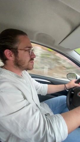 The video shows a man driving a car on a winding road. The man has brown hair and a beard, and he is wearing glasses, a light-colored shirt, and blue jeans. The car appears to be a Fiat. Inside the car, one can see the steering wheel, dashboard, rearview mirror, and a cross hanging from the rearview mirror. The road outside the car is bordered by greenery, and it is a cloudy day.