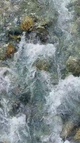 A top-down shot of a rocky riverbed with clear, turbulent water flowing over the stones. The water is churning and white with foam in some areas, indicating a fast current. The stones are of various sizes and colors, with some appearing smooth and others rough. Sunlight reflects off the water's surface, creating bright glints.