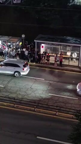The video, captured at night, shows a crowd of people gathered at a bus stop. The bus stop structure is visible, and a few cars are driving on the road in front of it. The people appear to be waiting for a bus or some form of transportation, with some individuals interacting amongst themselves. The lighting is artificial, suggesting it's evening or night. The overall environment appears to be urban or suburban.
