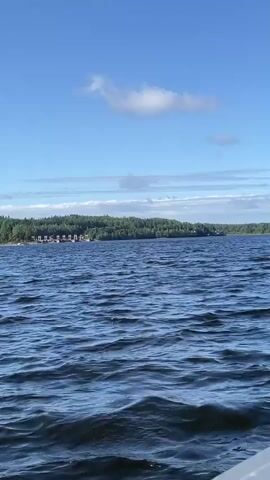 The video begins with a view of a calm blue lake under a bright blue sky with scattered white clouds. The shoreline, covered with dense green trees, is visible in the distance. As the video progresses, the camera pans to reveal the interior of a boat, showing the windshield, a steering wheel, seats, and various compartments. The boat appears to be moving, as indicated by the rippling water and the changing perspective. The overall scene is one of a peaceful day on the water, with the boat cruising along a picturesque lake surrounded by lush forest.