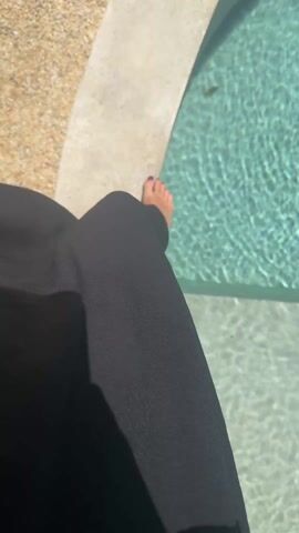 The video shows a small, round pool with clear turquoise water. The pool is bordered by a concrete edge and a gravelly area. On one side of the pool, a wooden platform extends over the water. Plants and a white decorative tree-like structure surround the pool. At the start of the video, a person wearing a black skirt stands at the edge of the pool, showing their bare feet with dark nail polish. The camera pans around the pool area.