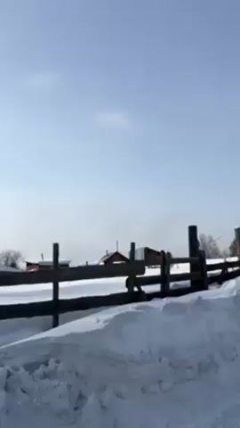 The video shows a snow-covered rural landscape on a bright, sunny day. A wooden fence and gate are visible in the foreground, partially obscured by snowdrifts. Beyond the fence, there are buildings in the distance and bare trees scattered throughout the scene. The camera appears to be moving, possibly from a vehicle passing by the fence.