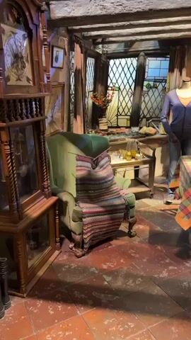 This video captures a scene from what appears to be a set, likely from a film or television production, in Watford, United Kingdom. The setting is an interior space, possibly a kitchen or dining area, with a rustic and somewhat cluttered aesthetic.

Dominating the left side of the frame is a tall, ornate wooden grandfather clock. To its right, a plush green armchair is draped with a colorful, hand-knitted blanket. Behind the armchair, a table with a checkered tablecloth is set for a meal, with plates, glasses, and a loaf of bread visible.

In the background, a mannequin dressed in a blue cardigan and jeans stands near a dresser filled with various dishes, cups, and decorative items. The room features exposed wooden beams on the ceiling and a large diamond-paned window with stained glass elements. The floor is tiled with reddish-brown terracotta tiles.

The scene is lit, suggesting it's an indoor, controlled environment, possibly a museum exhibit or a recreation of a historical or fantasy setting. There are no people actively present in the scene, only the mannequin. The overall impression is one of a cozy, lived-in space, though its staged nature is evident. No text is visible in the video.