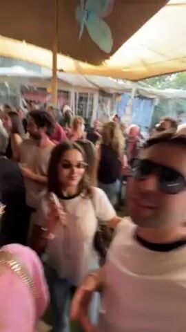 A man and a woman wearing sunglasses are at an outdoor party with a large crowd of people. They are taking a selfie video while dancing and smiling. The party seems festive, with colorful decorations hanging from the ceiling and music playing in the background. The crowd is dancing and enjoying the event.