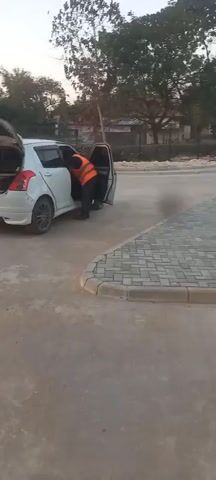 A person wearing an orange vest is seen near a white car with its trunk and driver's side door open. The person appears to be interacting with the inside of the car. The car is parked on a paved surface next to a curb made of brick pavers. Trees and buildings are visible in the background. The video appears to be shot during daylight.