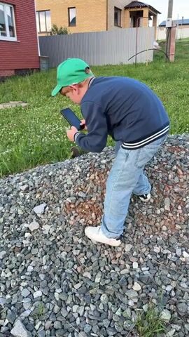 A young boy with red hair, wearing a green baseball cap, a dark blue jacket with white stripes, and light blue jeans, is engrossed in his smartphone. He is crouched down, looking intently at the screen while holding it in his right hand. His left hand is extended, possibly reaching towards something on the ground. He is standing on a pile of gravel.

In the background, a brick house with white trim and windows is visible. Adjacent to the house is a grassy area with scattered white flowers and some dirt. Further back, a fence can be seen, and beyond that, more houses. The overall scene suggests an outdoor setting, possibly a yard or a construction area, during daylight hours with fair weather. There's a slight incline in the terrain. A few small, dark objects that could be cats are visible in the grass behind the boy. The text "Maxi" is visible on the boy's green cap. The location context provided is Pyzhany, Russia. The boy's focused expression indicates deep engagement with his phone.