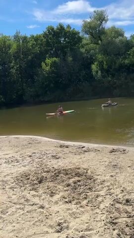 A wide shot shows a sandy beach in the foreground with the edge of a body of water in the middle ground. Two people are visible on the water in kayaks or canoes. The person in the foreground kayak is on the left side of the frame and is paddling. The person in the background kayak is on the right side of the frame and is also paddling. The water is a greenish-brown color. Dense green trees line the far bank of the body of water. The sky is blue with some white clouds. The overall impression is a sunny day at a natural body of water.