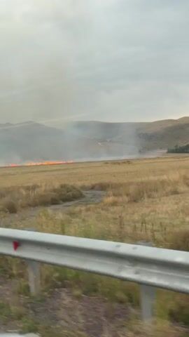 A landscape video captured from a moving vehicle shows a field of dry grass with a line of fire visible in the distance, producing smoke. The sky is overcast with clouds. In the foreground, a silver guardrail is visible, indicating the perspective is from a road. The terrain consists of rolling hills in the background.