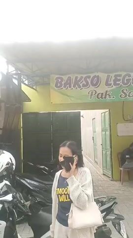 The video shows the exterior of a restaurant named BAKSO LEGENDA Pak So. A woman wearing a mask, a light-colored cardigan over a dark t-shirt, and white shorts is standing on the left side of the screen, holding a white purse and talking on a mobile phone. In the background, there are several motorcycles parked. The entrance to the restaurant is visible, with people inside sitting at tables. One man is sitting on a chair outside the restaurant, looking at his phone. Another man is walking towards the entrance, and a green motorcycle is parked in front of the entrance. A white car is parked on the right side of the screen. A banner above the entrance advertises a promotion for purchases on Fridays. The overall environment appears to be a casual dining establishment.