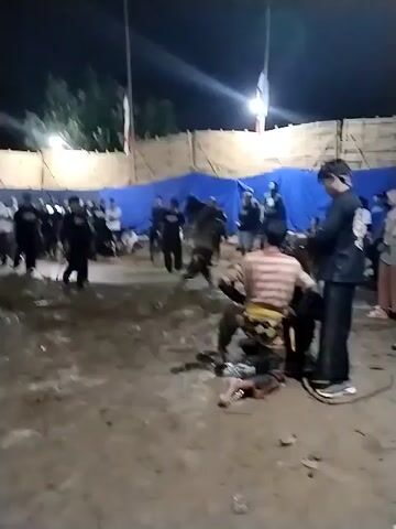 The video depicts an outdoor scene at night in Jenggrik, Indonesia. The ground is extremely muddy and wet, indicating recent rain. Artificial lights illuminate the area. A group of people, primarily men, are gathered. In the initial frame, a man in a striped shirt and patterned lower garment kneels on the muddy ground, while another man in a black shirt stands over him, holding a whip or braided rope. In later shots, a man actively swings a long whip, striking the muddy ground, causing splashes. Other individuals stand or move around in the background. Temporary structures, including blue tarpaulins and what appears to be a bamboo fence, are visible, along with a white building with a distinctive roof in the background. The activity involves whips and appears to be part of a traditional performance or ritual. No discernible text is visible.