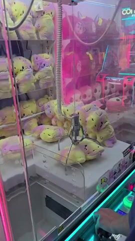 The video shows a claw machine filled with stuffed animals. The claw descends and attempts to grab a yellow and purple stuffed animal. The claw successfully grabs the animal and lifts it. The stuffed animal is then dropped into the prize chute. A hand is visible on the right side of the screen, operating the controls of the machine. The machine is brightly lit with neon pink and green lights. The background is filled with more stuffed animals, including some larger ones in pink and purple.