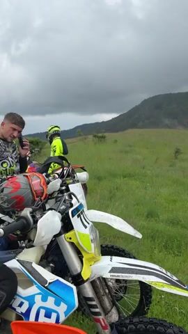 The video opens with an outdoor shot of a hilly landscape, featuring green grassy terrain in the foreground and a tree-covered mountain under an overcast sky. The camera pans right, revealing more distant mountains and a valley, with the sky gradually showing patches of blue. Two people in motorcycle gear and helmets are then shown standing next to a dirt bike on the grassy hill. The video concludes with a close-up shot of one of the individuals, wearing a black helmet with white accents, looking directly at the camera.