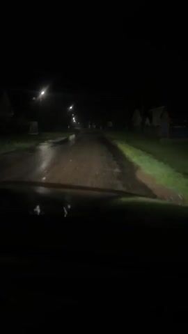 The video is taken from inside a vehicle driving on a dark, wet road at night. Streetlights illuminate parts of the road, which appears to be unpaved or poorly maintained, with puddles and uneven surfaces. Houses and vegetation are visible on either side of the road. The camera is pointed forward, showing the perspective of the driver. The overall impression is of a rural or semi-rural setting at night, possibly after rain.