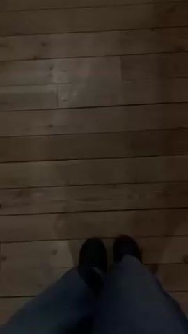 A first-person view shows a person wearing blue jeans and black shoes walking on a wooden floor. The floor is made of planks, and the video is slightly dark.