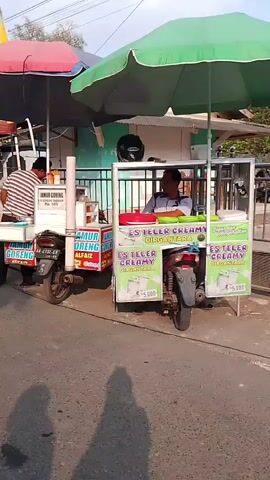 The video shows a street scene with several food stalls. In the foreground, there are two mobile food carts on motorcycles. The cart on the left is selling fried "jamur" (mushrooms) and "goreng" (fried food) labeled "LAMUR GORENG ALFAIZ". The cart on the right is selling "ES TELOR CREAMY" (creamy egg ice) labeled "DIRGANTARA". A man is visible behind the ES TELOR cart, wearing a white and blue striped shirt. To the left of the jamur goreng cart, another man is visible wearing a striped shirt and dark pants, appearing to be preparing food. Behind these carts, there is a longer, more permanent stall with a red canopy, displaying various food items and snacks for sale. This stall also has pictures of food items along its front. The ground is paved, and shadows suggest it is daytime with sunlight. The overall environment appears to be a street market or roadside food vendor area.