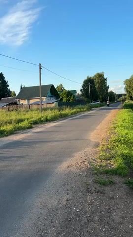 A person rides a motorcycle down a paved road through a rural village. The motorcycle is moving towards the viewer. The rider is wearing a light-colored shirt and dark pants. The road is bordered by green grass and trees. In the background, there are houses with pitched roofs. The sky is blue with a few white clouds. The sun appears to be setting, casting long shadows.