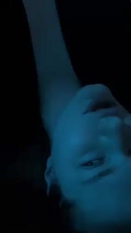 A close-up, dimly lit shot of a person's face, looking upwards. The lighting gives the face a blue hue, and the person's eyes are partially open. The background is dark.  There's what appears to be a raised arm visible in the upper left. The person's expression isn't readily discernible due to the lighting and angle. There is music playing in the background.