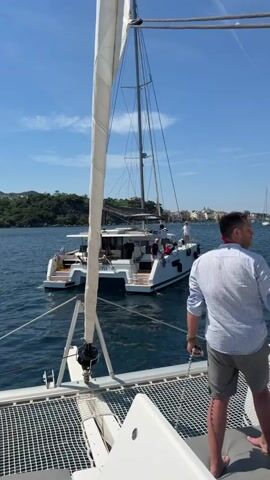 A man, viewed from behind, stands on the deck of a white catamaran sailboat. He is wearing a light blue, striped button-down shirt and gray shorts. He is holding a metal chain attached to the boat. The deck of the catamaran is made of a net material. In the background, another white catamaran sailboat with passengers on board is visible. The name "Latona" is printed on the back of the catamaran. The sky is blue and clear with some clouds. Green hills can be seen in the distance. The water is a dark blue. The man speaks and an engine sound is heard.