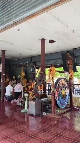 Here is a description of the video:

The video shows the interior of what appears to be a Buddhist shrine or temple. Two people are kneeling in prayer before a row of golden Buddha statues. A figure of the elephant-headed deity Ganesha is prominently displayed, adorned with orange garlands. A large gong, decorated with colorful patterns, stands nearby. A woman in traditional clothing walks into frame and appears to be preparing to play a drum. The floor is covered with red tile, and the room is open-air with a corrugated metal roof overhead.