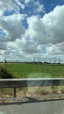 The video shows a view from a moving vehicle looking out at a field of green crops. In the distance, a long line of towering electrical pylons is visible against a sky filled with white, puffy clouds. The pylons are a dark grey color and are spaced evenly apart, supporting thick electrical cables. The field is lush and green, with rows of crops extending towards the pylons. A metal guardrail runs along the bottom of the frame, suggesting the vantage point is from a road or highway. The sun is shining, casting shadows and illuminating the scene. The overall impression is one of rural industrial landscape.