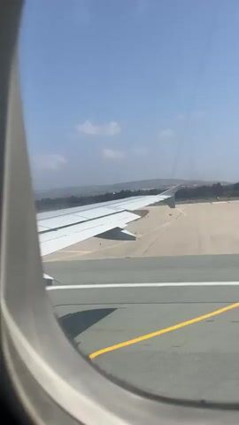 The video shows a view from an airplane window, looking out at a runway. The airplane's wing is visible in the foreground, taking up a significant portion of the frame. The runway extends into the distance, with a brown, dry landscape surrounding it. The sky is blue with a few scattered clouds. The airplane appears to be either taking off or landing, as it is close to the ground. There are no people visible in the video. The overall impression is one of movement and travel.