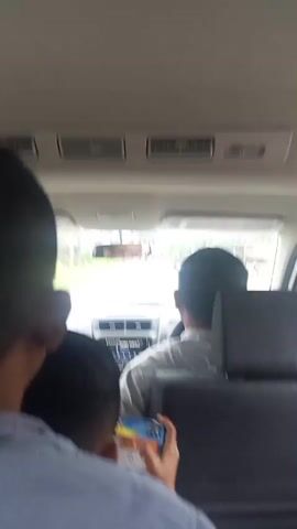 The video is filmed from the back seat of a car, looking forward. In the foreground, a young person's head is visible, looking down at a smartphone. Behind them, another young person's head is visible. In the middle ground, a person with dark hair is driving the car. The dashboard and front windshield are visible, showing a blurry outdoor scene. In the background, through the side windows, a landscape of green fields and structures can be seen, moving past the car. The video appears to capture a moment of travel, with one person using a phone and others observing the journey.