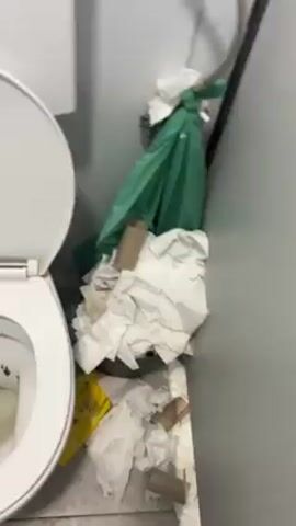 Here's a forensic description of the scene depicted in the video:

**Scene:** A dirty, unsanitary bathroom.

**Objects:**
*   A white toilet with the lid open. The toilet bowl contains discolored liquid and visible fecal matter.
*   Toilet paper scraps and empty toilet paper rolls scattered around the toilet and on the floor.
*   A green garbage bag visible partially behind the toilet.
*   Brown stains, possibly fecal matter, smeared across the tiled floor.
*   A pair of athletic shoes worn by the videographer in the lower part of the frame.

**Overall Impression:** The scene indicates a poorly maintained and unhygienic bathroom environment. There is evidence of human waste and a lack of cleaning.