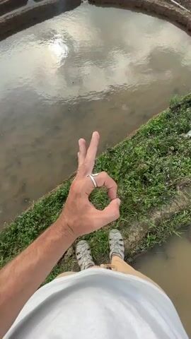 The video begins with a low-angle shot of a person's hand, adorned with a ring, making an "OK" gesture in the foreground. The person's sandals and lower attire are also visible. Below the hand, a terraced rice paddy filled with water reflects the cloudy sky and a bright light source, likely the sun. Green grass and small plants grow on the edge of the paddy.

As the camera slowly pans upwards and slightly to the right, the hand remains in the bottom left corner, gradually revealing a vast landscape of meticulously carved terraced rice fields. These fields stretch across multiple hillsides and into the distance, characteristic of mountainous regions like Lao Chai, Vietnam. Many of the paddies are filled with water, creating shimmering reflections of the dramatic, cloud-filled sky, with patches of bright sunlight breaking through. Other sections of the terraces are dry, showing the rich, dark earth. The expansive vista of intricate terraces against the backdrop of distant mountains under a dynamic sky creates a serene and impressive scene, suggesting a late afternoon or early evening setting.