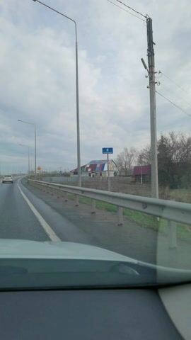 A vehicle is driving down a wet highway on an overcast day. Street lights are on either side of the road, and a white car is driving ahead. A building with multicolored squares on its roof is visible on the right. A yield sign is visible on the side of the road. A guardrail is on the right of the road.