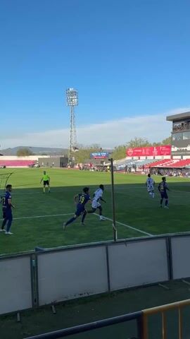The video shows a soccer game in progress on a sunny day. Players in white and blue uniforms compete on a green field, striving to score against the opposing team. A goalkeeper in pink stands ready to defend the goal, while other players are positioned around the field. The scene is observed from behind a barrier, with a stadium and surrounding landscape visible in the background, including a tall lighting tower and various buildings. Additionally, a security guard wearing a yellow vest and a red beret stands nearby, facing the field.