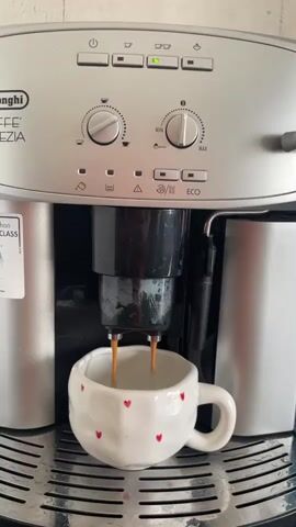 A silver Delonghi coffee machine is dispensing two streams of dark coffee into a white mug with red hearts on a silver grate. The machine has two dials and several buttons. A sticker is visible on the left side of the machine.