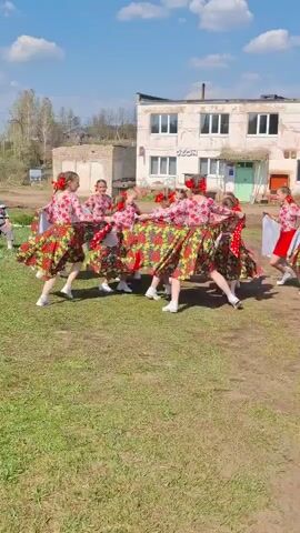 A group of young girls dressed in traditional Russian attire are performing a folk dance in a circle on a grassy field. They are wearing colorful, floral-patterned dresses with red accents and white shoes. Their hair is styled with red ribbons or flowers. The girls are holding hands and moving in a coordinated fashion. In the background, there is a two-story building with an "OZON" sign visible, suggesting it might be a store or office. The sky is blue with scattered clouds, indicating a sunny day. The surrounding area appears to be a rural or small-town setting.