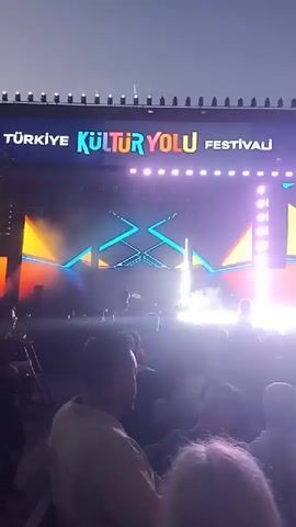 The video shows a large crowd of people gathered in front of a brightly lit stage at an outdoor event. The stage features the words "Türkiye Kültür Yolu Festivali" displayed in colorful lettering, along with the words "Turizm Bakanliği." The stage design incorporates geometric shapes and is illuminated with a variety of lights, including spotlights and LED panels. There is a significant amount of haze or fog surrounding the stage, adding to the visual effect. The crowd is dense, with people of various ages and styles visible. The atmosphere appears lively and energetic, suggesting a music performance or other type of entertainment. The sky is visible above the stage, suggesting it is either dusk or night.