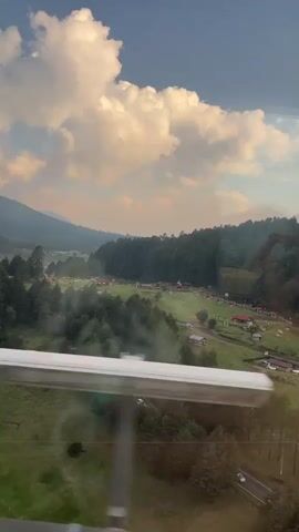 The video shows a landscape featuring a mix of forests, meadows, and low-lying buildings, likely taken from an elevated perspective, possibly from inside a vehicle in motion. The scene is dominated by green vegetation, with dense forests extending across much of the frame. Interspersed among the trees are open meadows and clearings with small buildings, many with red roofs. In the background, mountain ranges are visible, partially obscured by haze. The sky overhead is blue with scattered white clouds. The overall lighting suggests it's either early morning or late afternoon, with soft, diffused sunlight bathing the scene. The movement indicates the shot is likely taken from a moving vehicle.  At the bottom of the frame, part of a metal rail is visible.