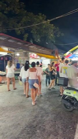 The video shows a nighttime outdoor market scene. Several people are walking and standing around stalls. The stalls appear to be selling seafood, with tanks of live seafood visible. The market is lit by artificial lights from the stalls and overhead. Trees are visible in the background. The ground is covered in gravel. The overall atmosphere appears to be bustling and lively.