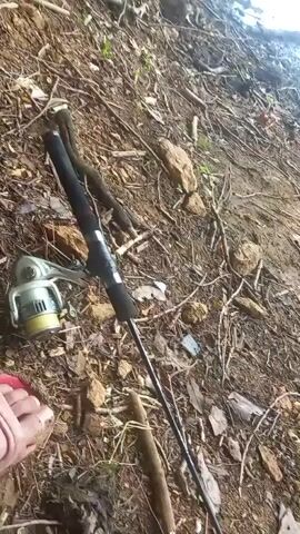 The video begins with a close-up of a fishing rod resting on the muddy bank of a body of water. The camera pans to reveal a scenic view of the lake, with trees and rolling hills in the background. A person's hand enters the frame, holding a lit cigarette, and they take a drag. The camera then focuses on the hand and cigarette as the person exhales smoke. The video ends with a shot of the lake and surrounding landscape.