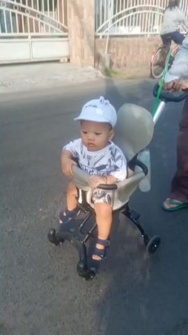 A baby wearing a white cap and a white t-shirt with a black and white pattern sits in a stroller. The baby is looking forward with a neutral expression. The stroller is on a paved surface, likely a sidewalk or street. In the background, a white fence is visible. A person's legs and feet, wearing a brown garment and sandals, are partially visible to the right of the stroller, suggesting they are pushing it. The sun is casting shadows.