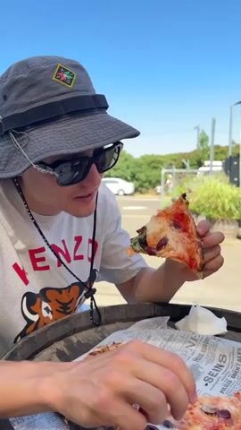 The video shows a close-up of a person wearing a grey bucket hat and sunglasses, eating a slice of pizza outdoors. The person is holding the pizza with their right hand and taking a bite. They are wearing a white t-shirt with a red graphic and a black lanyard around their neck. The pizza is on a newspaper, which is placed on a wooden surface, possibly a barrel. The background is slightly blurred but shows greenery, a building, and a clear blue sky, suggesting a sunny day. The time of day appears to be daytime, likely late morning or afternoon due to the bright sunlight. The setting appears to be casual, possibly a restaurant or outdoor seating area. The person's focus is on eating the pizza, and their expression is neutral. Visible text includes "KENZO" on the person's t-shirt and text on the newspaper, which is mostly illegible but appears to be in French, with words like "SEINS" and "ORIENTALES" being partially visible. The location context provided is Golan Regional Council, Israel.