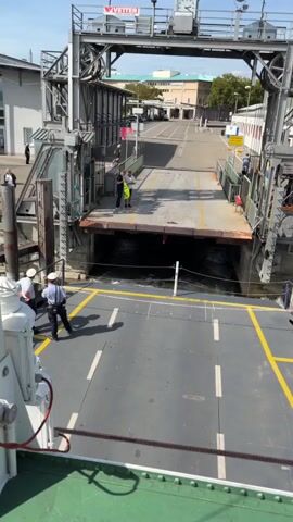 A ferry is docked at a port. The ferry's ramp is lowered and connects to the shore, creating a gateway for vehicles. The water below the ramp is choppy and turbulent. Several people, possibly crew members or port workers, are standing on the ferry's deck near the ramp, dressed in uniforms or work attire. In the background, there is a building and a paved area that appears to be a road or parking lot. The sky is clear and the day is sunny.