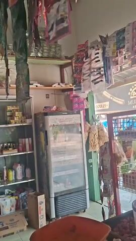 The video pans across the interior of a small store, likely a convenience store or minimart. The shelves are filled with various products, including packaged snacks, beverages, toiletries, and what appear to be cleaning supplies. There are also several boxes on the floor. A refrigerator unit is visible, stocked with items. Some products are hanging from the ceiling. The lighting is somewhat dim. The store appears to be well-stocked but cluttered. No people are visible in the video.